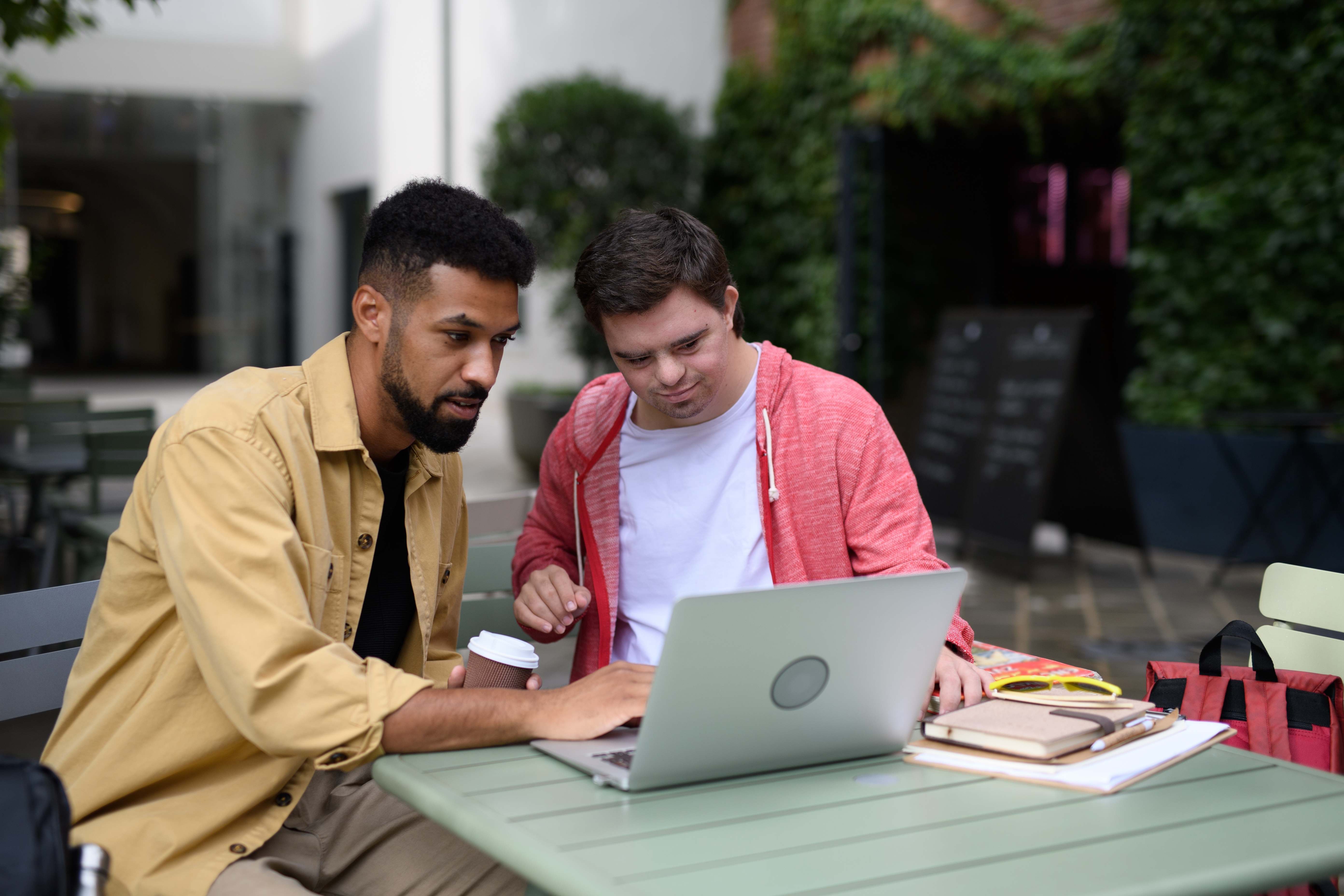 Caregiver helping young man with Down syndrome learn computer skills at outdoor cafe