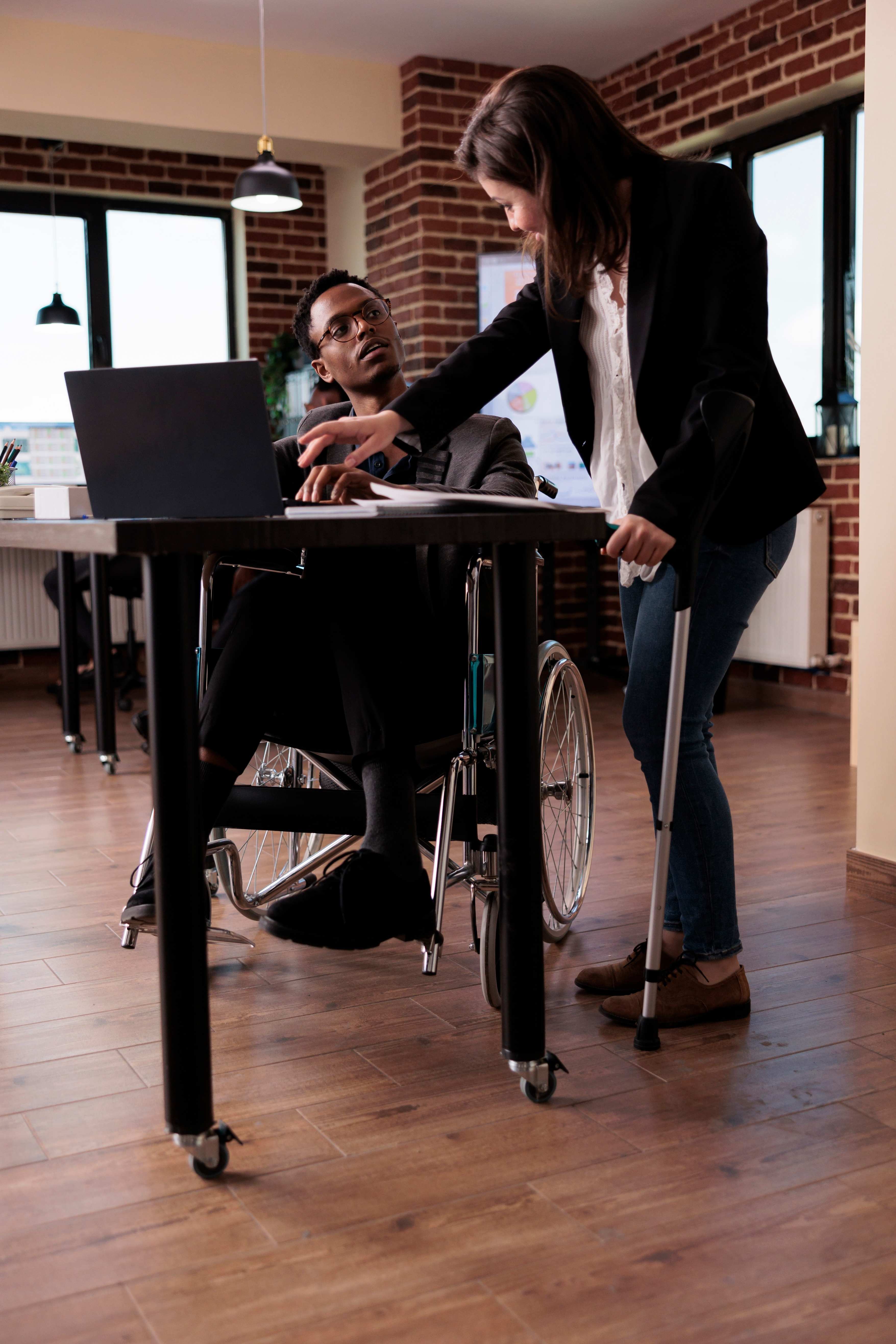 Man in wheelchair working on laptop with colleague support in office